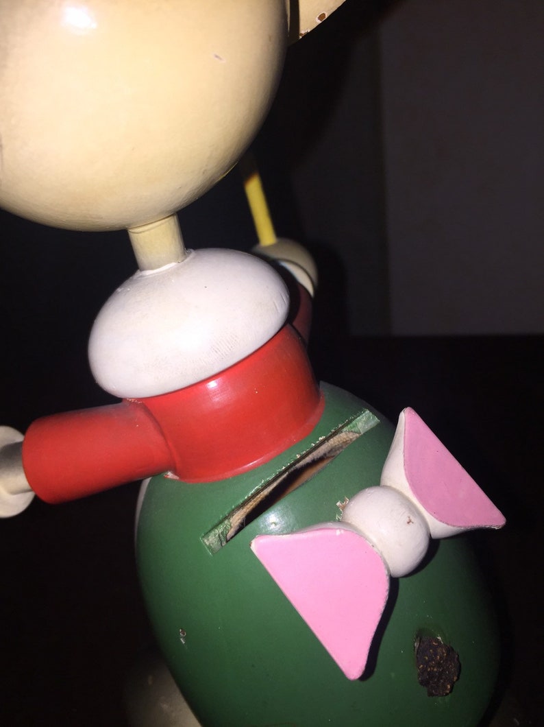 以下が含まれることがあります： 大きな頭と緑色の体を持つフィギュアの形をした、ヴィンテージの木製おもちゃの銀行。フィギュアは赤いスカーフ、白い帽子、ピンクのリボンをしています。銀行にはコインを挿入するためのスロットがあります。