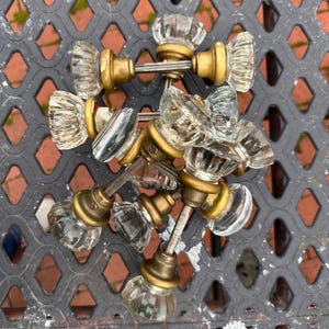 May include: A collection of clear glass door knobs with brass-colored bases and stems. The knobs have a faceted, rounded design. The hardware is arranged on a dark gray metal surface with a diamond pattern.