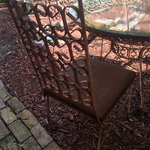 May include: A rusty metal patio chair with a brown cushion. The chair is made of intricate metalwork and has a unique design. The chair is sitting on a bed of brown wood chips.