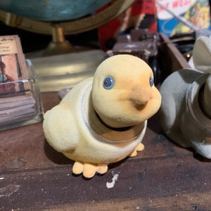 May include: A vintage yellow plush duck toy with blue button eyes and a silver collar. The duck has a rounded body and small feet. The toy is sitting on a dark wooden surface.