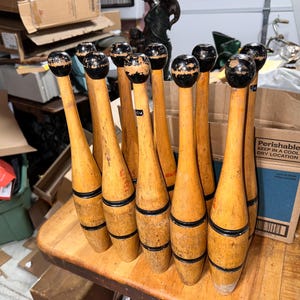 May include: A collection of ten wooden exercise clubs with a light brown finish, each featuring a rounded black top and two black horizontal bands around the midsection. The clubs are arranged upright in a cluster, showing signs of wear.