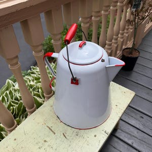 May include: White enamelware kettle with red accents. The kettle has a red handle and a red trim around the lid and spout. The kettle is sitting on a weathered, light yellow wooden surface.