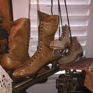 May include: A pair of brown leather ice skates with laces and metal blades. The skates are hanging from hooks on a white wooden wall.