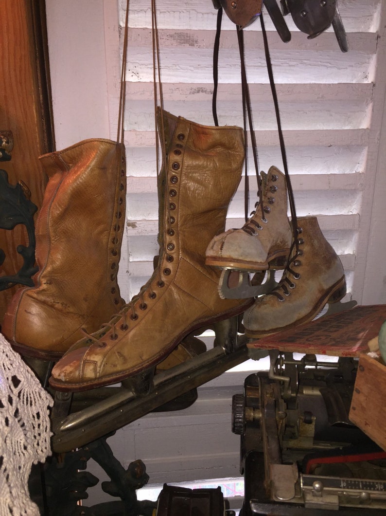 May include: A pair of brown leather ice skates with laces and metal blades. The skates are hanging from hooks on a white wooden wall.