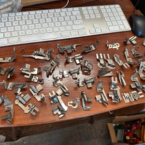 May include: A collection of silver sewing machine feet, including a variety of styles and sizes, laid out on a wooden surface.