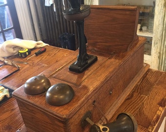 May include: Antique wooden telephone with a black receiver, two brass bells, and a black rotary dial. The phone is sitting on a wooden table.