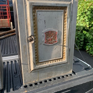May include: A vintage metal cabinet with a red and gold label that reads "The Original Pop Corn Machine". The cabinet is gray and has a decorative border around the door.