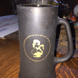 May include: Black ceramic mug with a gold Playboy Club logo featuring a silhouette of a woman holding a key. The logo is surrounded by the text "The Playboy Club" repeated multiple times.