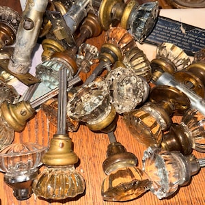 May include: A collection of antique door knobs in various styles and materials. The knobs feature clear glass, brass, and metal components. Some have faceted designs, while others are smooth. The assortment is arranged on a wooden surface.