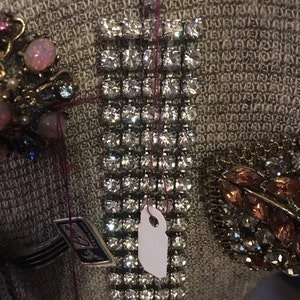 May include: A silver rhinestone bracelet with a white tag attached. The bracelet is made of small, square rhinestones set in a metal frame. The tag is blank and has a hole punched in the top.