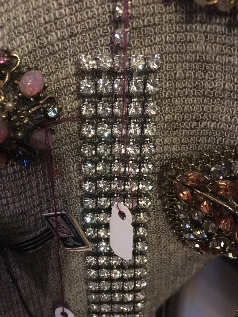 May include: A silver rhinestone bracelet with a white tag attached. The bracelet is made of small, square rhinestones set in a metal frame. The tag is blank and has a hole punched in the top.