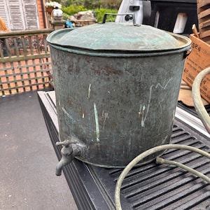 May include: A large, aged copper water container with a lid and a spigot. The container has a weathered, greenish-blue patina, and a handle on the lid. A coiled hose is visible near the container.