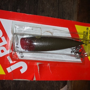 May include: A brown and red fishing lure with a silver hook, packaged in a clear plastic blister pack. The packaging is labeled "J-PLUG" and "LUHR JENSEN & SONS/HOOD RIVER OREGON".