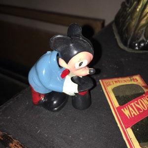 May include: A black and white wind-up toy of Mickey Mouse wearing a blue jacket and red pants. The toy is in a bent-over position, as if it is about to pick something up.