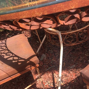 May include: A rusty metal patio set with a glass table top and four chairs. The chairs have a scrolled design and are made of white metal. The set is sitting on a bed of red wood chips.