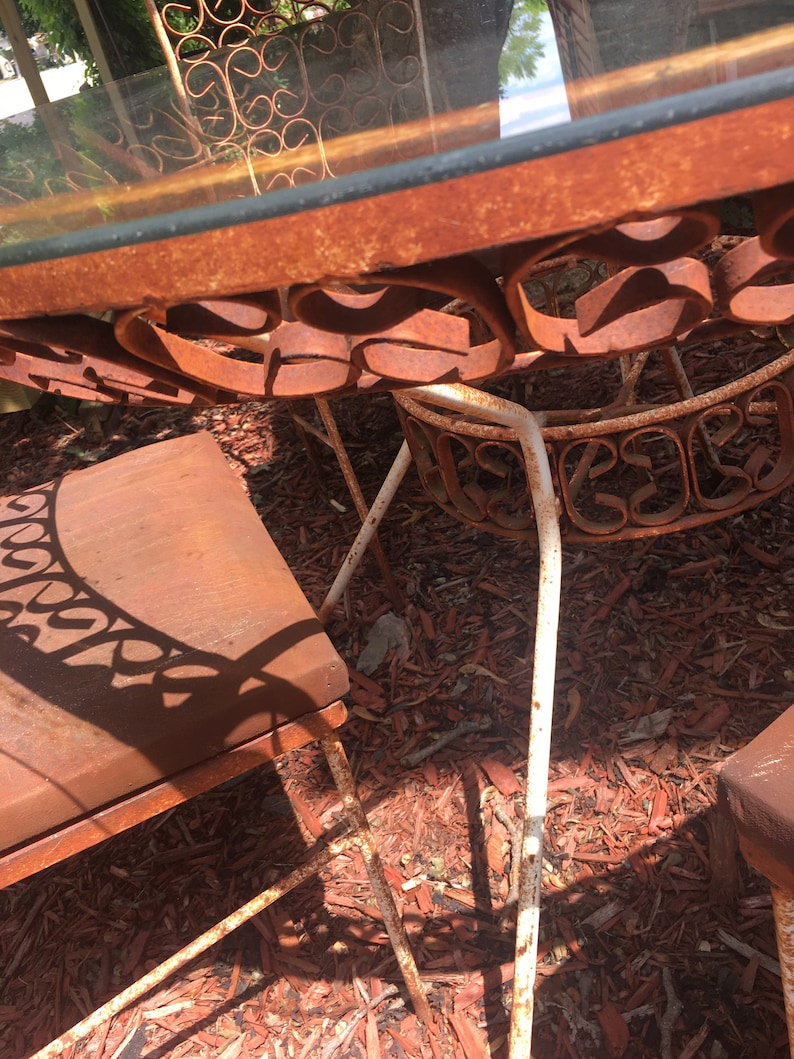 May include: A rusty metal patio set with a glass table top and four chairs. The chairs have a scrolled design and are made of white metal. The set is sitting on a bed of red wood chips.