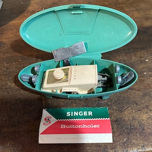 May include: A vintage Singer buttonholer in a teal green plastic case with a white buttonholer and a gold knob. The case is open and the buttonholer is visible. The case has a metal plate on the inside. The buttonholer is labeled "Singer" and has a small label that says "Automatic". The buttonholer is accompanied by a white and red instruction card that says "Singer Buttonholer The Singer Company".