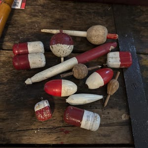 May include: A collection of vintage fishing floats in various shapes and sizes, featuring red and white painted finishes. These wooden and cork bobbers include cylindrical, spherical, and tapered designs, some showing signs of wear and weathered textures.