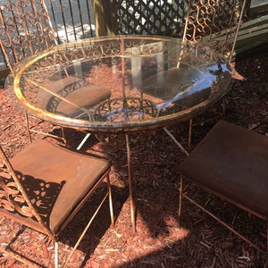 May include: A rusty metal patio set with a round glass table top and four chairs. The chairs have ornate metalwork and brown cushions. The set is sitting on a bed of red wood chips.