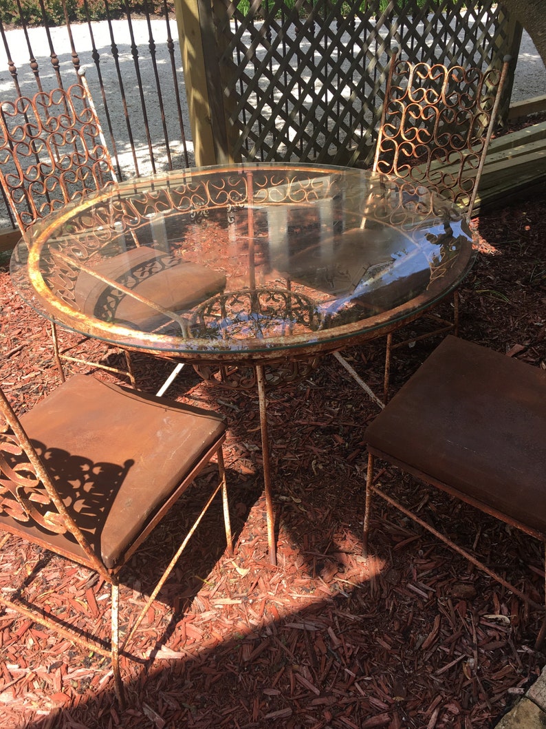 May include: A rusty metal patio set with a round glass table top and four chairs. The chairs have ornate metalwork and brown cushions. The set is sitting on a bed of red wood chips.