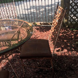 May include: A set of four rusty metal chairs and a glass-top table with a decorative metal base. The chairs and table are sitting on a bed of red wood chips.