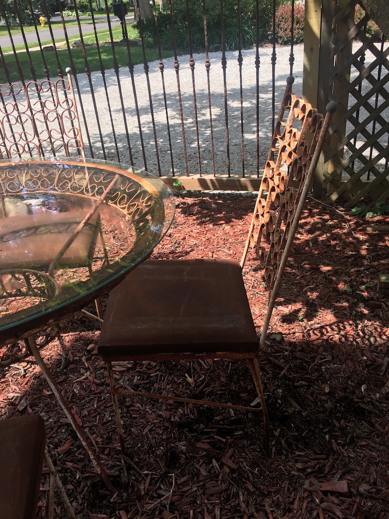 May include: A set of four rusty metal chairs and a glass-top table with a decorative metal base. The chairs and table are sitting on a bed of red wood chips.