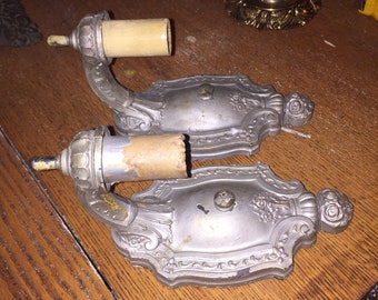 May include: Two antique silver wall sconces with ornate designs and a decorative scrollwork pattern. Each sconce has a ceramic light bulb socket and a brown paper lampshade.
