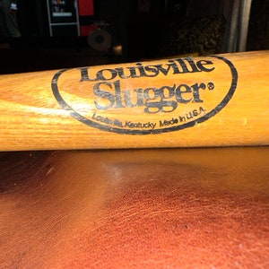 May include: A wooden Louisville Slugger baseball bat with a black oval logo that reads "Louisville Slugger" and "Louisville Kentucky Made in U.S.A."