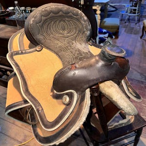 May include: A brown leather Western saddle with decorative stitching and a tan canvas seat. The saddle features a dark brown leather seat with a detailed stitched design and is resting on a wooden chair.