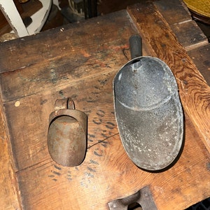 May include: Two vintage metal scoops, one with a handle and one without, on a wooden surface. The scoops are both made of metal and have a worn, rustic look.
