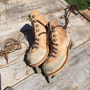May include: A pair of vintage ice skates with brown leather boots and silver blades. The laces are tied and the skates are sitting on a wooden surface.