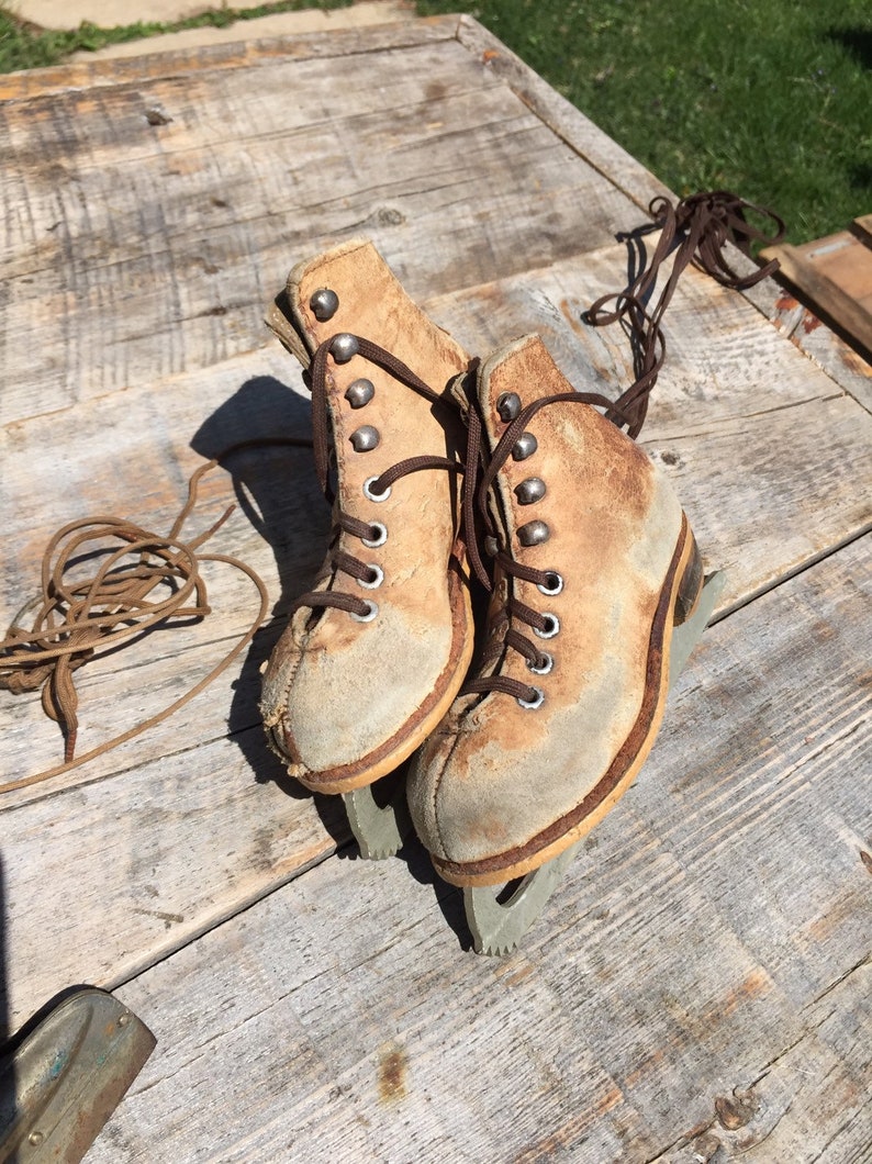 May include: A pair of vintage ice skates with brown leather boots and silver blades. The laces are tied and the skates are sitting on a wooden surface.
