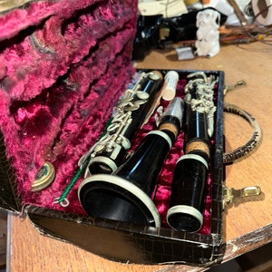 May include: A black clarinet with silver keys and a black case with a red velvet interior. The clarinet is in the case with a cleaning rod and a cork grease tube.