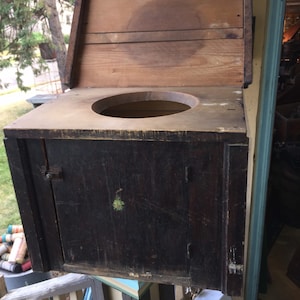 May include: A wooden outhouse with a hinged lid and a small door. The outhouse is painted black with a green stain on the front. The lid is open and the inside of the outhouse is visible.