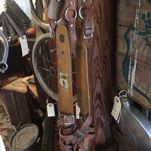 May include: Vintage wooden skis with leather bindings and metal buckles. The skis are a light brown color and show signs of wear. A red wooden handle is visible at the top. The image also includes a bicycle wheel and other antique items.