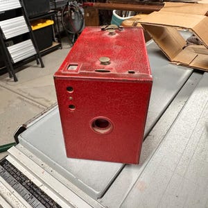 May include: A vintage red box camera with a textured surface. The camera has a rectangular shape with a circular lens opening and several smaller holes. It features brass-colored hardware and a classic design.