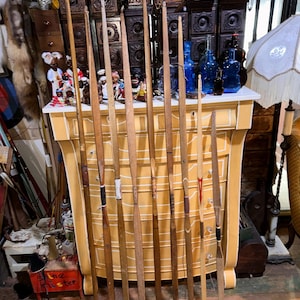 May include: A collection of eight long wooden archery bows standing vertically in a row. The bows vary in wood tone from light to dark brown, with some featuring leather grips, string notches, and decorative accents in black, white, and red.
