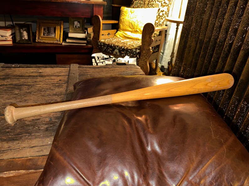 May include: A wooden baseball bat with a natural finish. The bat is lying on a brown leather surface.