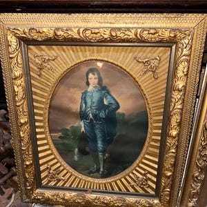 May include: A framed portrait of a young boy wearing a blue suit and holding a hat. The portrait is set against a background of a landscape with trees and a river. The frame is ornate and gold.