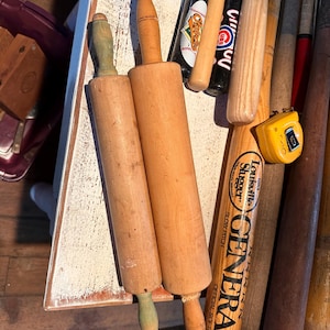 K&ouml;nnte beinhalten: Zwei h&ouml;lzerne Nudelrollen mit hellbraunen Walzen und gedrehten Griffen, eine mit gr&uuml;nem Griff. Mehrere Baseballschl&auml;ger, darunter einer mit "Louisville Slugger" und "GENERAL" aufgedruckt, und ein gelbes Ma&szlig;band sind ebenfalls zu sehen.