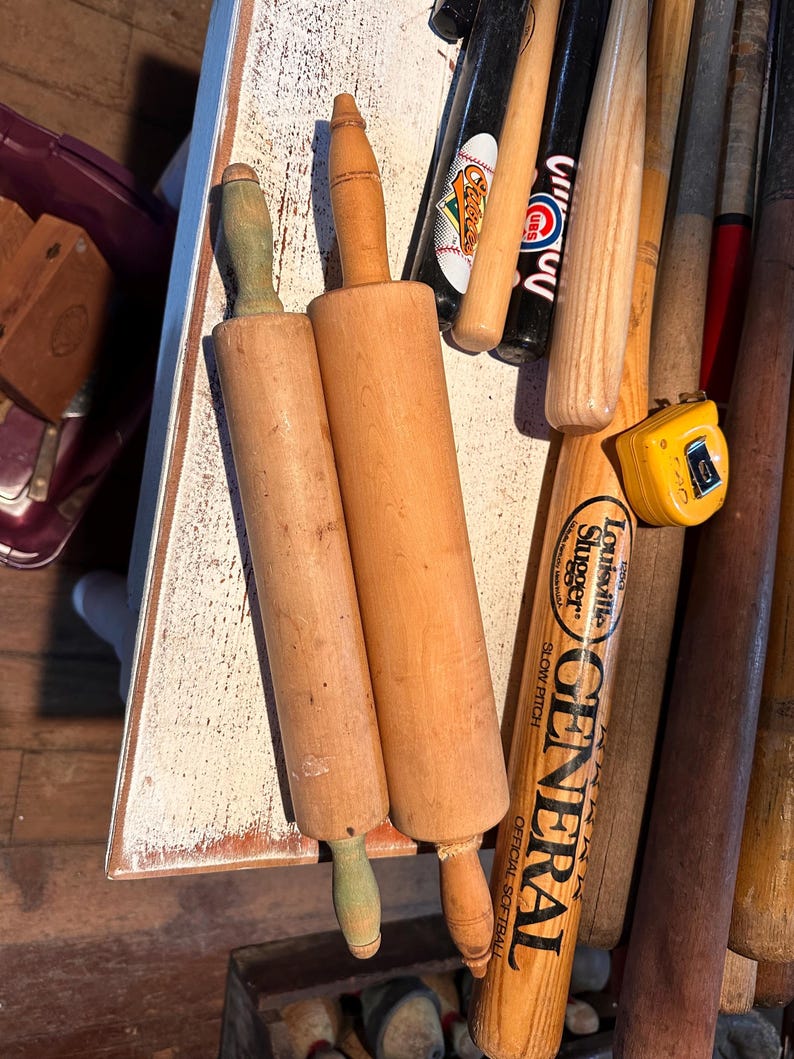 K&ouml;nnte beinhalten: Zwei h&ouml;lzerne Nudelrollen mit hellbraunen Walzen und gedrehten Griffen, eine mit gr&uuml;nem Griff. Mehrere Baseballschl&auml;ger, darunter einer mit "Louisville Slugger" und "GENERAL" aufgedruckt, und ein gelbes Ma&szlig;band sind ebenfalls zu sehen.