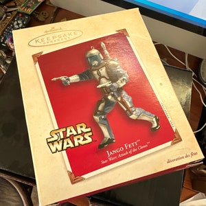 May include: A red and white Hallmark Keepsake Ornament featuring Jango Fett from Star Wars: Attack of the Clones. The ornament depicts Jango Fett in his Mandalorian armor, holding a blaster rifle and a jetpack.
