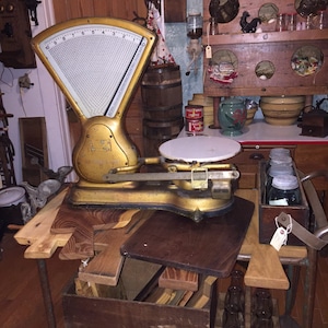 May include: A vintage gold-colored scale with a white dial and a black platform. The scale is sitting on a wooden table with a variety of other items, including a wooden box, a wooden crate, and a collection of glass bottles.