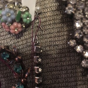May include: A close-up of a brown fabric with a variety of jewelry pieces, including a floral brooch with pink, blue, and green stones, a silver chain with clear stones, and a silver chain with a pearl.