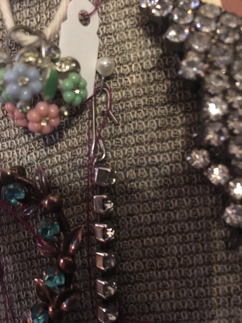May include: A close-up of a brown fabric with a variety of jewelry pieces, including a floral brooch with pink, blue, and green stones, a silver chain with clear stones, and a silver chain with a pearl.