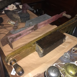 May include: Two antique wooden tools with a hand crank and a metal handle. The tools are on a brown paper surface. The tool on the left has a metal handle and a red painted surface. The tool on the right has a wooden handle and a brown painted surface. The tools are in a vintage shop setting.