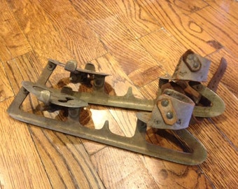 May include: A pair of vintage ice skates with leather straps. The skates are made of metal and have a rusty patina. The straps are worn and have holes in them.