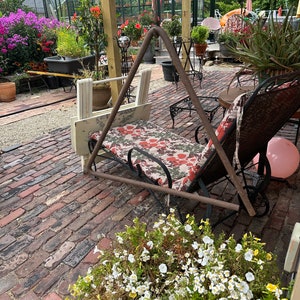 May include: A metal swing hanging from a wooden pergola in a brick patio. The swing is made of a triangular frame with a cushioned seat. There are flowers and plants in pots around the patio.
