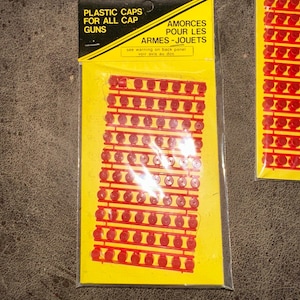 May include: Two packages of plastic cap gun ammunition. The packaging is yellow with black text that reads "PLASTIC CAPS FOR ALL CAP GUNS" and "AMORCES POUR LES ARMES-JOUETS". The caps are red and arranged in rows.