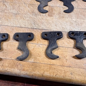 May include: Five black metal hooks with a T-shaped base and a curved hook. The hooks are on a wooden surface.